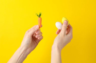 Sarı arka planda plastik havuç ve renkli yumurta tutan el. Paskalya konsepti