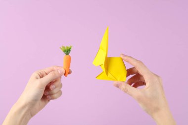 Mor arka planda plastik havuç ve origami tavşanı tutan bir el. Paskalya konsepti