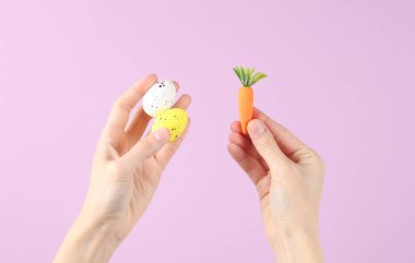 Mor arka planda plastik havuç ve renkli yumurta tutan bir el. Paskalya konsepti