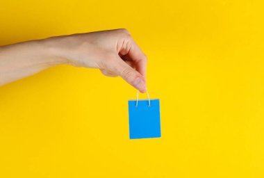 Kadının eli sarı arka planda mavi minyatür alışveriş çantası tutuyor. Satış, alışveriş konsepti