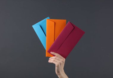 Kadının eli koyu arkaplanda üç renkli zarf tutuyor.