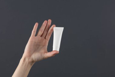 Kadının eli koyu gri arka planda beyaz krema tüpünü tutuyor. Tasarım için şablon, yaratıcı güzellik modeli