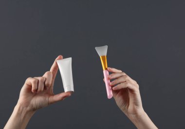 Kadının eli, koyu gri arka planda beyaz krem tüpü ve plastik fırçayı tutuyor. Tasarım için şablon, yaratıcı güzellik modeli