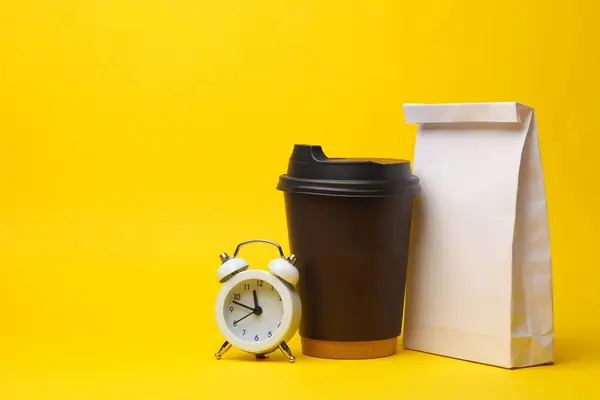 Paper lunch bags with cardboard coffee cup and alarm clock on yellow background