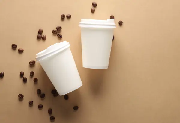 White Disposable cardboard cups with coffee beans on brown beige background. Creative layout. Top view