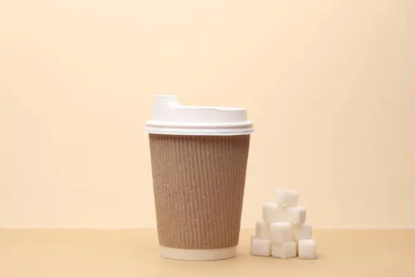 Disposable coffee cup and sugar cubes on beige background
