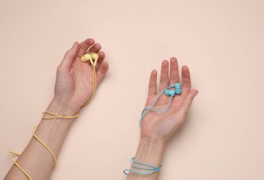 Kadın eli, bej arka planda mavi ve sarı kablolu kulaklıkları tutar.