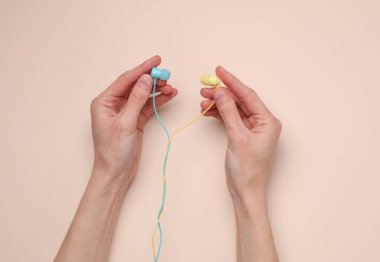 Kadın eli, bej arka planda mavi ve sarı kablolu kulaklıkları tutar.