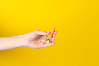 Sarı arka planda plastik minyatür havuç tutan bir el.