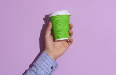 Mor pastel arka planda yeşil karton kahve fincanı tutan bir adam.