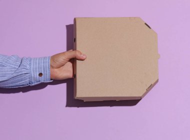 Mor pastel arka planda, üzerinde gölgesi olan karton pizza kutusu tutan bir adam..