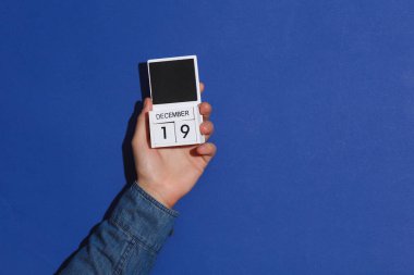 Kot gömlekli adamın elinde ahşap blok takvim düzenleyicisi var. Tarihi 19 Aralık, mavi arka planda.