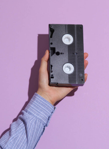 Мужская рука в рубашке с видеокассетой на фиолетовом пастельном фоне с тенью