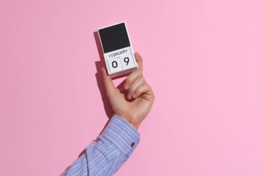 9 Şubat tarihli ahşap blok takvim düzenleyicisi, pembe arka planda, gölgeli bir adamın elini tutuyor.