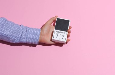 Adamın eli gömlekli, tahta bloklu takvim düzenleyicisi 11 Şubat 'ta pembe arka planda, gölgeli.