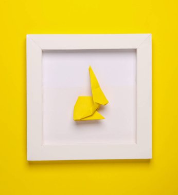 Origami tavşanı beyaz kare çerçevede, sarı arka planda. Estetik asgari natürmort. Kavramsal fotoğraf. Üst görünüm