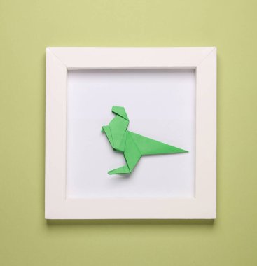 Origami dinozoru beyaz kare çerçevede, yeşil arka planda. Estetik asgari natürmort. Kavramsal fotoğraf. Modern sanat. Üst görünüm