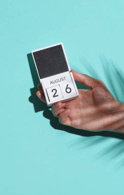 Bir adamın elinde tahta blok takvimi var. 26 Ağustos 'ta mavi arka planda, palmiye yaprağı gölgesinde.