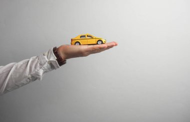 Beyaz tişörtlü adamın elinde gri arka planda oyuncak taksi var.