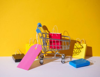 Minyatür kağıt alışveriş torbalarıyla süpermarket arabası ve sarı gri arka planda satış etiketi. Kara Cuma, dünya alışveriş günü, tatil indirimi konsepti.