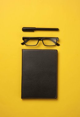 Siyah deri kapaklı, kalemli, sarı arka planda gözlüklü defter. İş konsepti. Üst Manzara. Düz yatıyordu