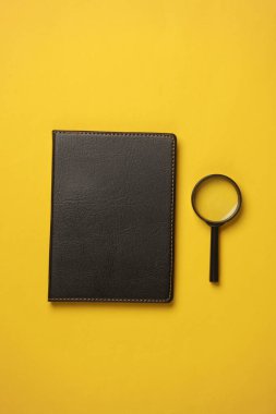 Siyah deri kaplı defter ve sarı arka planda büyüteç.