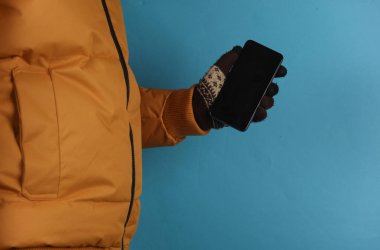 Sarı ceketli ve eldivenli bir adam mavi arka planda akıllı telefon tutuyor.