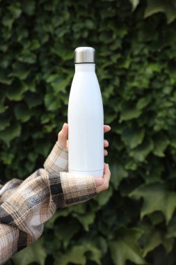 Elleri beyaz termos şişesini yeşil yaprak arka planında tutuyor.