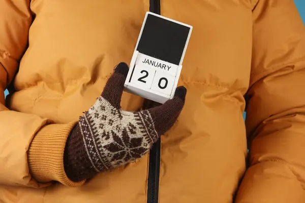 stock image Man in yellow winter down jacket and gloves holding block calendar with date january 20 on blue background