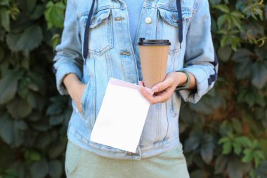 Elinde beyaz kağıt beslenme çantası ve yeşil bitkilerin arka planına karşı kahve fincanı tutan kadın.