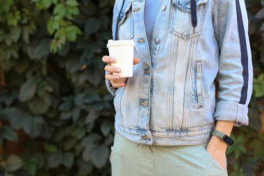 Kot ceketli bir kadın yeşil bitki arka planında karton kahve fincanı tutuyor.