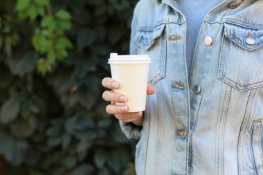 Kot ceketli bir kadın yeşil bitki arka planında karton kahve fincanı tutuyor.