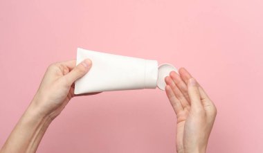 Pembe arka planda beyaz krema tüpü tutan kadın eller.
