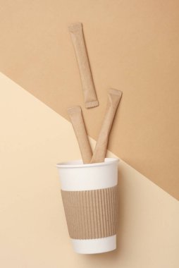 Sıcak içecekler için tek kullanımlık karton bardak ve bej arka planda şekerli veya kahveli çubuklar.