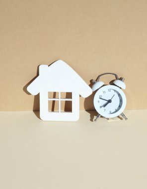 Beyaz ahşap ev modeli ve gölgeli bej bir arka planda çalar saat.