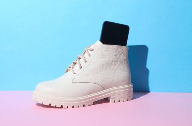 Pembe mavi arka planda gölgeli akıllı telefonlu beyaz kadın deri çizmesi. Yaratıcı düzen. Minimalizm modadır.