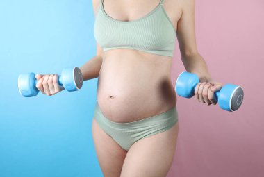 İç çamaşırlı hamile bir kadın mavi pembe arka planda dambıllar tutuyor. Hamilelik boyunca egzersiz.