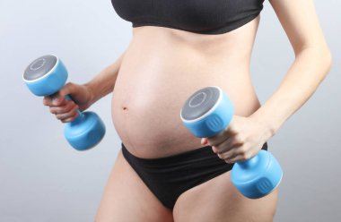 İç çamaşırı eğitimi almış, elinde dambıllarla gri arka planda hamile bir kadın. Hamilelik boyunca egzersiz.