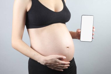 Elinde gri arka planda beyaz bir ekranla akıllı telefon tutan hamile bir kadın. Model, tasarım şablonu, bilgileriniz