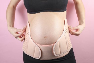 Sırtı ve göbeği için ortopedik kemeri olan hamile bir kadın. Hamile kadınlar için bandaj. Üçüncü üç aylık dönem için özel kemer. Pembe arka planda stüdyo çekimi