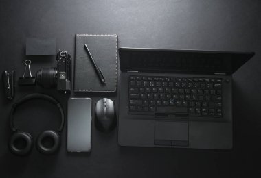 Modern siyah aletler ve aksesuarlar. Laptop, kamera, akıllı telefon, stereo kulaklık, siyah arka planda not defteri. Çalışma alanı. Düz yatıyordu. Üst görünüm