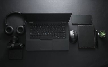 Modern siyah aletler ve aksesuarlar. Laptop, kamera, akıllı telefon, stereo kulaklık, siyah arka planda not defteri. Çalışma alanı. Düz yatıyordu. Üst görünüm