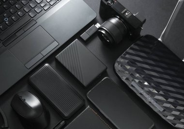 Modern siyah cihazlar ve aksesuarlar, laptop, kamera, akıllı telefon, harici sabit disk, kablosuz ağ yönlendirici, USB flash disk, siyah arka planda güç bankası. Fotoğrafçı ekipmanı. Çalışma alanı. Düz yatıyordu. Üst görünüm