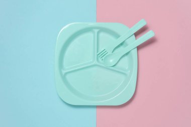 Pembe mavi arka planda bebeği beslemek için kaşık ve çatalla ayrı plastik tabak. Üst görünüm