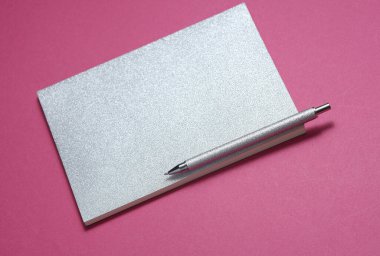 Gümüş kapaklı not defteri ve pembe arka planda kalem.