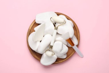 Masaj silindiri ve yüz masajı için gua sha kazıyıcı, vücut yağı spatulası, pembe arka planda çakıl taşları. Üst görünüm