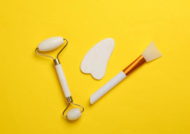 Yeşim yüz silindiri ve gua sha kazıyıcı, sarı arka planda vücut yağı spatulası. Güzellik konsepti. Üst görünüm