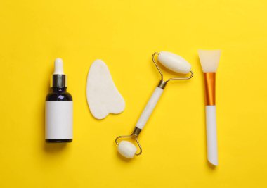 Yeşim yüz silindiri ve gua sha kazıyıcı, serum şişesi, sarı arka planda vücut yağı spatulası. Güzellik konsepti. Üst görünüm