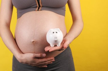 Sarı arka planda karnını ve kumbarasını tutan hamile bir kadın.