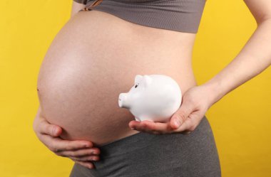 Sarı arka planda karnını ve kumbarasını tutan hamile bir kadın.